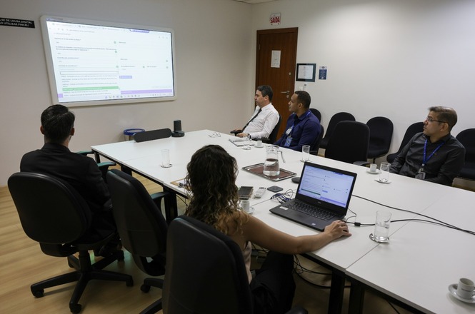 Sala de reunião com cinco pessoas sentadas ao redor de uma mesa retangular. Na mesa, há um laptop aberto em frente a uma mulher de cabelos cacheados, que está de costas. O laptop exibe uma tela semelhante à projetada na parede à frente, onde há um projetor mostrando um documento ou apresentação. As outras quatro pessoas, três homens e uma mulher, estão sentadas ao redor da mesa, olhando para a projeção. Há copos de vidro e xícaras de café sobre a mesa. A sala tem piso de madeira clara e uma porta de madeira ao fundo, com uma placa de "Saída" acima.