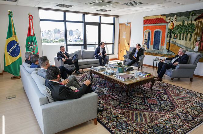 Foto colorida: autoridades sentadas em sofás, sala institucional com vista urbana, reunião oficial, debatendo pautas em bege, azul e verde