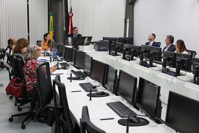 Sala de conferência com várias pessoas sentadas em torno de uma mesa em formato de "U". Na parte interna da mesa, há vários computadores e monitores. À esquerda, algumas pessoas estão sentadas, incluindo uma mulher de cabelos grisalhos e blusa vermelha estampada. À direita, há três homens e uma mulher, todos em trajes formais. No fundo, há uma bandeira do Brasil e uma bandeira vermelha com detalhes em verde e dourado. As paredes são brancas com persianas verticais. O ambiente parece ser um espaço de trabalho ou reunião formal.