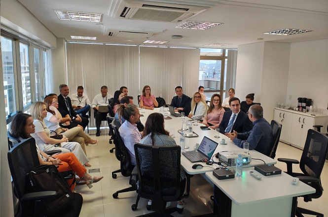 Sala de reunião com várias pessoas sentadas ao redor de uma mesa grande. A maioria das pessoas está vestida formalmente, com ternos e roupas sociais. Há um projetor e um laptop sobre a mesa, sugerindo que uma apresentação ou discussão está em andamento. A sala tem janelas grandes com persianas fechadas, e há um armário ao fundo com garrafas de café e copos. As pessoas parecem estar atentas e engajadas na conversa.