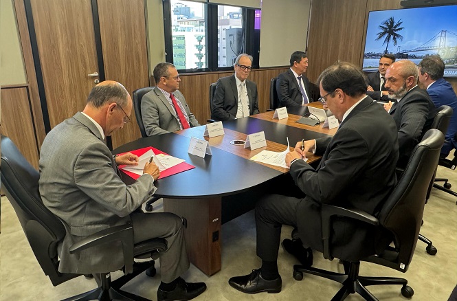 Sala de reunião com sete homens de terno sentados ao redor de uma mesa oval de madeira. Eles estão envolvidos em uma discussão ou assinatura de documentos. Há placas de identificação na mesa em frente a cada pessoa. A sala possui paredes de madeira e janelas ao fundo, através das quais é possível ver prédios. À direita, há uma tela grande com uma imagem de uma ponte e a palavra "FLORIANÓPOLIS". O ambiente parece formal e corporativo.
