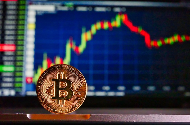 Uma moeda de Bitcoin em destaque na frente de uma tela que mostra um gráfico de velas, comum em análises de mercado financeiro. O gráfico tem velas verdes e vermelhas, indicando variações no preço ao longo do tempo. A moeda tem o símbolo do Bitcoin, um "B" com duas linhas verticais. A imagem sugere um contexto de negociação ou investimento em criptomoedas.