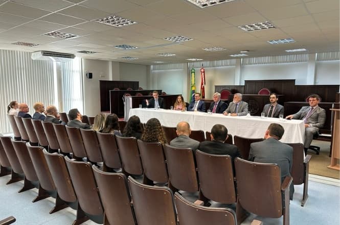 Sala de conferências ou tribunal com várias fileiras de cadeiras de madeira voltadas para uma mesa comprida coberta com uma toalha branca. Atrás da mesa, há oito pessoas sentadas, todas vestidas formalmente com ternos e gravatas. Na parede ao fundo, há bandeiras, incluindo a bandeira do Brasil. O ambiente é iluminado por luzes de teto e há janelas com persianas verticais ao longo da parede lateral. Na frente das cadeiras, há um púlpito de madeira.