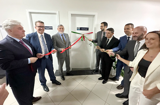 Um grupo de nove pessoas está participando de uma cerimônia de inauguração. Elas estão de pé em frente a duas portas brancas, segurando uma fita vermelha e verde que está prestes a ser cortada. Todos estão vestidos formalmente, com ternos e roupas sociais. As portas têm placas azuis com nomes escritos nelas. A atmosfera parece ser de celebração e formalidade.