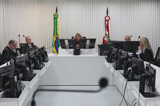 A imagem mostra uma sala de tribunal com cinco pessoas sentadas atrás de mesas em formato de U, cada uma com monitores de computador à sua frente. Duas bandeiras, uma do Brasil e outra de um estado, estão posicionadas ao fundo. As pessoas parecem ser juízes ou advogados, vestindo trajes formais. A cena sugere uma sessão de julgamento ou audiência jurídica.