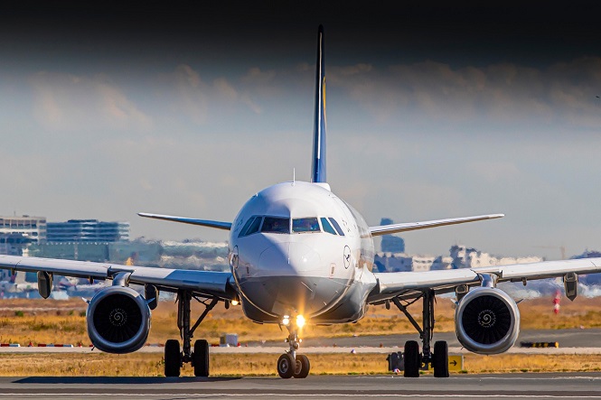 A imagem mostra um avião em uma pista de aeroporto, visto de frente. O avião é um modelo de jato comercial, e podemos ver claramente suas duas asas e motores. Ao fundo, há uma paisagem urbana com alguns edifícios, e o céu está parcialmente nublado. A luz do sol ilumina a cena, destacando o avião no centro da imagem.