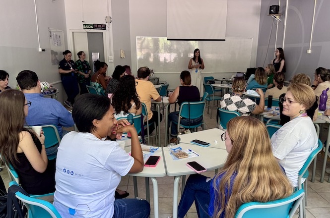A imagem mostra uma sala de aula com várias pessoas sentadas em carteiras dispostas em grupos. Elas parecem estar participando de uma apresentação ou palestra. Uma mulher está de pé na frente, falando para o grupo, enquanto outra está de pé mais ao lado, aparentemente auxiliando na apresentação. Há lousas brancas ao fundo e o ambiente possui iluminação natural. As pessoas estão atentas, algumas com notebooks, celulares e garrafas de água sobre as mesas. A sala parece ser um local de educação ou treinamento.