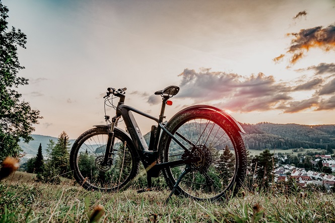 A imagem mostra uma bicicleta em uma área de grama, com um cenário de montanhas e árvores ao fundo. O céu está parcialmente nublado com tons alaranjados, indicando possivelmente um pôr do sol. Ao fundo, é possível ver uma vila ou cidade pequena, com várias casas. As cores são vibrantes, destacando a cena ao ar livre.