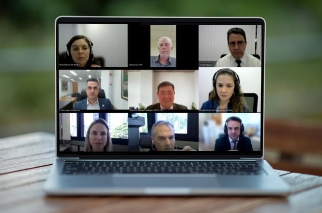 A imagem mostra uma videoconferência em um laptop. Na tela, há nove pessoas, cada uma em seu próprio quadrado, participando de uma reunião online. Algumas usam fones de ouvido. O ambiente parece ser formal, sugerindo uma reunião de negócios ou acadêmica. O laptop está sobre uma superfície de madeira, e o fundo é desfocado, dando a entender que o computador está em um ambiente natural ou iluminado.