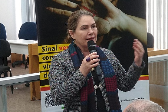 Na imagem, uma mulher está falando ao microfone em frente a um banner. Ela veste um casaco cinza e um cachecol colorido. No fundo, o banner parece estar relacionado a uma campanha contra a violência doméstica, com mãos e a frase "Sinal vermelho" visível. O ambiente parece ser uma sala ou auditório com móveis e cadeiras.