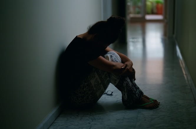 A imagem mostra uma pessoa sentada no chão de um corredor sombrio. A pessoa está encostada na parede, com os braços em volta dos joelhos, e parece estar em uma posição reflexiva ou preocupada. O ambiente é pouco iluminado, com luz ao fundo, criando uma atmosfera introspectiva