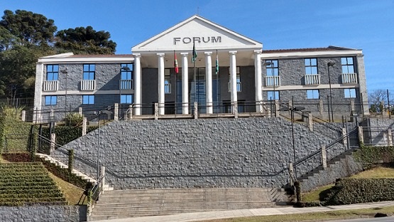 A imagem mostra um edifício identificado como "FÓRUM" na fachada. O prédio tem um design neoclássico, com colunas brancas na entrada principal, que é precedida por uma escadaria ampla. O edifício é simétrico, com duas alas laterais e várias janelas. Na frente, há bandeiras hasteadas. A estrutura está situada em uma área ajardinada com arbustos e grama ao redor.