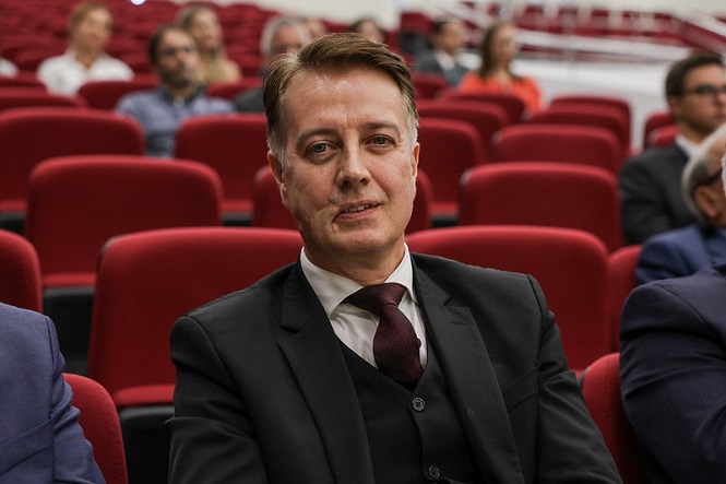 A imagem mostra um homem sentado em uma audiência ou conferência, usando um terno escuro com camisa branca e gravata. Ele está em uma fileira de poltronas vermelhas, e há outras pessoas sentadas ao fundo, desfocadas. O ambiente parece ser uma sala de eventos ou auditório.