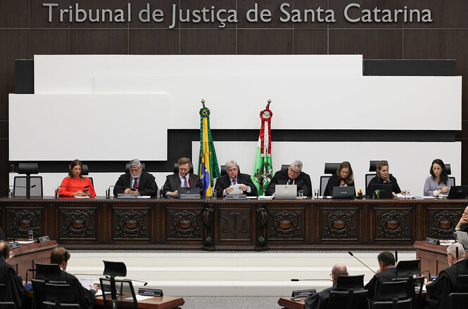 A imagem mostra uma sessão no Tribunal de Justiça de Santa Catarina. Há um grupo de pessoas sentadas em uma mesa longa, aparentemente juízes ou desembargadores, com bandeiras do Brasil e de Santa Catarina ao fundo. A mesa possui detalhes ornamentados em madeira e, ao fundo, está o nome do tribunal na parede. Várias pessoas estão trabalhando ou observando, e o ambiente é formal, típico de um tribunal.