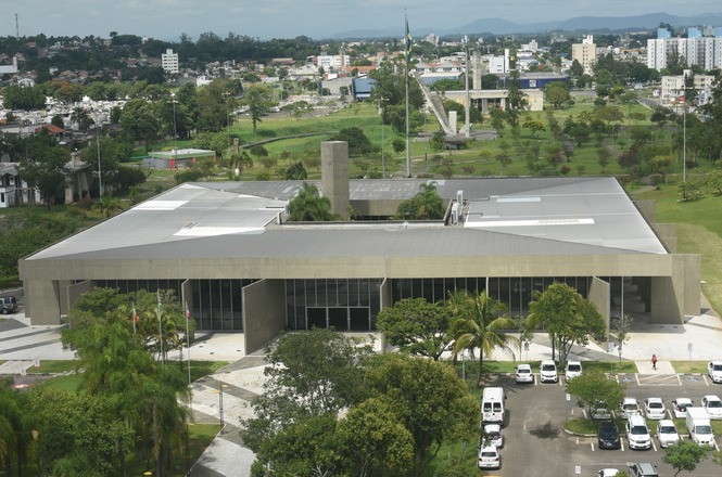 A imagem mostra um grande edifício moderno cercado por áreas verdes e vegetação. O prédio tem um design arquitetônico contemporâneo, com linhas retas e uma fachada de concreto aparente. Em frente ao edifício, há um estacionamento com vários carros. Ao fundo, é possível ver uma área urbana com prédios e árvores, além de um céu claro. A bandeira do Brasil está hasteada em um mastro à frente do edifício.