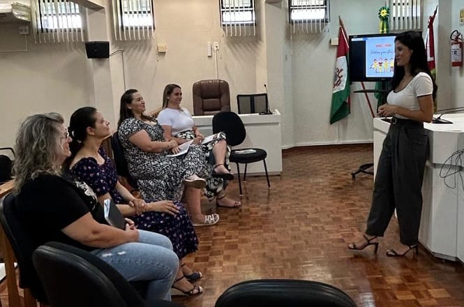 A imagem mostra uma sala de reunião com cinco mulheres. Quatro delas estão sentadas em cadeiras, enquanto uma está em pé falando para o grupo. À direita, há uma tela com uma apresentação e uma bandeira na parede. O chão é de madeira e o ambiente se assemelha a uma sala de conferências ou de aula.
