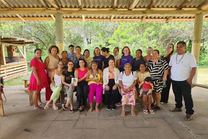 A imagem mostra um grupo de aproximadamente 23 pessoas reunidas sob uma estrutura coberta com telhado de zinco e pilares de concreto. O ambiente é cercado por árvores e vegetação, sugerindo uma área rural ou comunitária ao ar livre.
