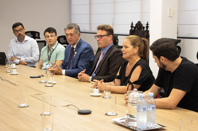Seis pessoas estão sentadas ao redor de uma mesa de madeira em uma sala de reunião. Da esquerda para a direita, há um homem de óculos com uma camisa branca, um homem de óculos com uma camisa verde clara, um homem de terno azul, um homem de terno escuro e óculos, uma mulher de cabelo preso e blusa preta, e um homem de barba com uma camiseta preta. Na mesa, há copos de vidro, garrafas de água mineral, xícaras de café e um controle remoto. Ao fundo, há cadeiras e uma janela com persianas fechadas.