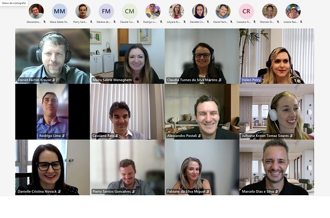 A imagem mostra uma videoconferência com várias pessoas. Há 15 participantes, todos aparecendo em janelas individuais. As pessoas estão sorrindo e parecem estar em um ambiente profissional ou de trabalho. Algumas usam fones de ouvido. Na parte superior, há uma barra com os ícones e iniciais dos participantes na chamada. O fundo das janelas varia de acordo com o ambiente de cada pessoa.