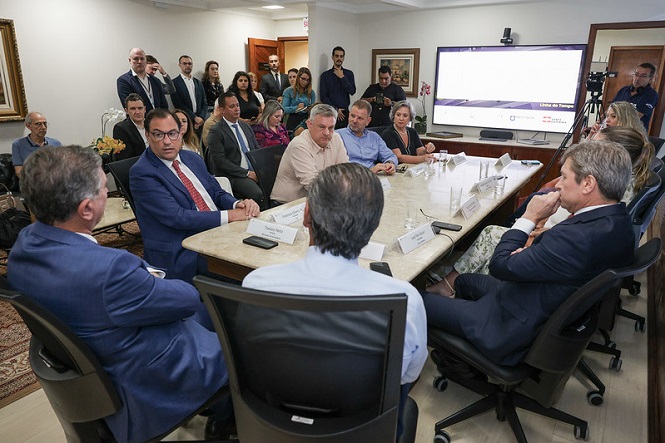 Sala de reuniões com várias pessoas sentadas ao redor de uma mesa retangular de mármore. Há homens e mulheres, alguns em trajes formais, como ternos e gravatas. No centro da mesa, há placas de identificação e microfones. Ao fundo, um grupo de pessoas está em pé, observando a reunião. Na parede, há uma grande tela de apresentação com gráficos e textos. O ambiente parece ser um escritório ou uma sala de conferências. Há também um fotógrafo ou cinegrafista no canto direito, capturando o evento.
