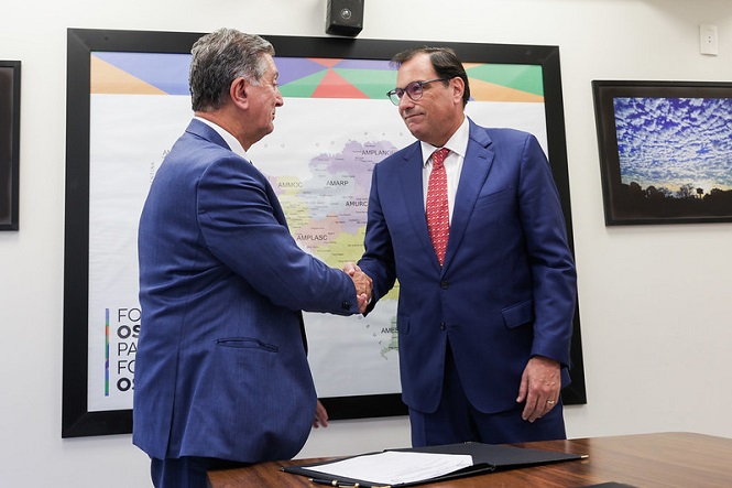 Dois homens de terno azul estão apertando as mãos em frente a um mapa emoldurado na parede. O homem à esquerda tem cabelo grisalho e está de perfil, enquanto o homem à direita usa óculos e uma gravata vermelha com bolinhas brancas. Na mesa à frente deles, há um documento aberto. Ao fundo, além do mapa, há uma fotografia de um céu azul com nuvens emoldurada na parede.