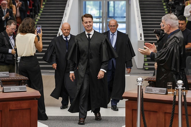 Um grupo de homens usando togas pretas está caminhando em direção à câmera em um ambiente formal, possivelmente um tribunal ou uma cerimônia oficial. O homem no centro parece ser o foco principal, caminhando com confiança. Atrás dele, há dois homens também em togas. À esquerda, uma mulher está tirando uma foto com o celular. À direita, um homem está aplaudindo. O ambiente é elegante, com móveis de madeira e uma escadaria ao fundo. Há outras pessoas ao redor, algumas aplaudindo e outras observando.