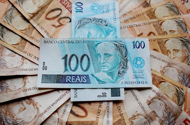 Notas de dinheiro brasileiro espalhadas, com destaque para uma nota de 100 reais no centro. Ao fundo, há várias notas de 50 reais. As notas de 100 reais são predominantemente azuis e apresentam a efígie da República, enquanto as notas de 50 reais são de cor marrom e também apresentam a mesma efígie.