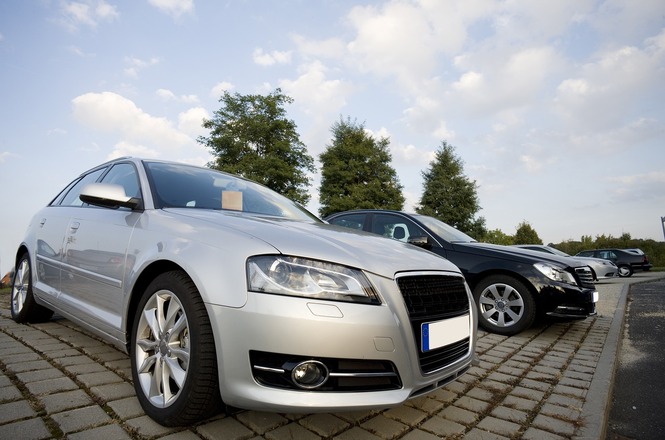 Dois carros estacionados em um pavimento de blocos de concreto. O carro em primeiro plano é um Audi prata, com design moderno e esportivo. Ao lado, há um carro preto, também com um design elegante. Ao fundo, algumas árvores e um céu parcialmente nublado. A cena parece ser em um estacionamento ao ar livre.