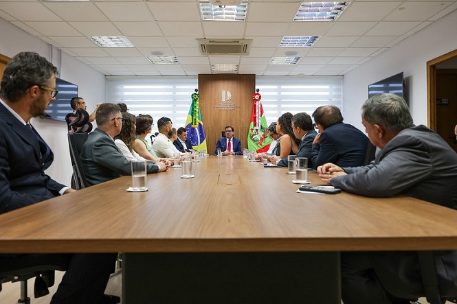 Foto horizontal: entre as bandeiras do Brasil e de SC, Francisco Oliveira Neto conduz reunião com servidores, mesa longa, tons claros.