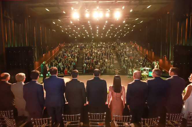 Foto colorida: autoridades no palco frente a grande público, auditório amplo iluminado, solenidade institucional, acompanhando cerimônia em dourado, preto e verde