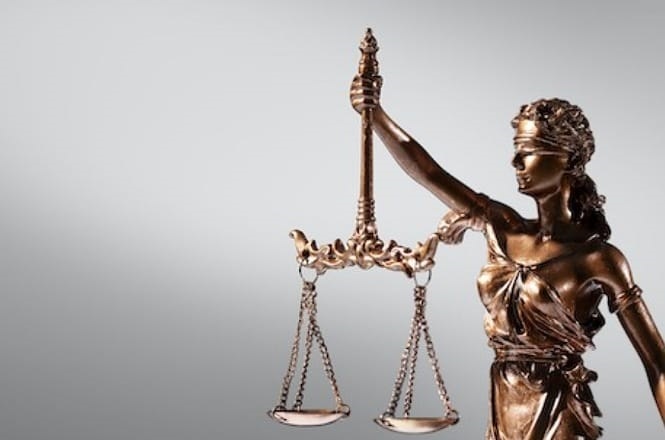 Estátua da deusa da justiça, conhecida como Lady Justice. Ela está vendada, simbolizando imparcialidade, e segura uma balança em uma das mãos, representando a justiça equilibrada. A estátua é feita de bronze e tem um acabamento brilhante. O fundo é simples e de cor cinza, destacando a figura da estátua.