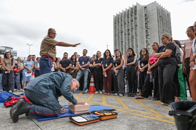 Um grupo de pessoas está reunido ao ar livre, assistindo a uma demonstração de primeiros socorros. No centro, um homem de uniforme azul está ajoelhado no chão, realizando compressões torácicas em um manequim de treinamento. Ao lado dele, há um instrutor de pé, gesticulando com a mão, provavelmente explicando o procedimento. O grupo de espectadores está atento, formando um semicírculo ao redor dos instrutores. No chão, há equipamentos de primeiros socorros dispostos ao lado do manequim. Ao fundo, é possível ver um edifício alto e moderno. O ambiente parece ser um espaço público ou estacionamento.