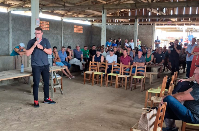 Um homem está em pé, falando ao microfone, em um espaço coberto com teto de madeira e paredes de concreto. Ele está usando uma camisa cinza escura e calças pretas, com tênis preto e vermelho. À sua frente, há várias fileiras de cadeiras de madeira, onde muitas pessoas estão sentadas, prestando atenção. Algumas pessoas estão sentadas em bancos de madeira ao fundo. O ambiente parece ser simples, com chão de terra batida e iluminação natural entrando pelas aberturas nas paredes. Há uma sensação de reunião comunitária ou evento local.