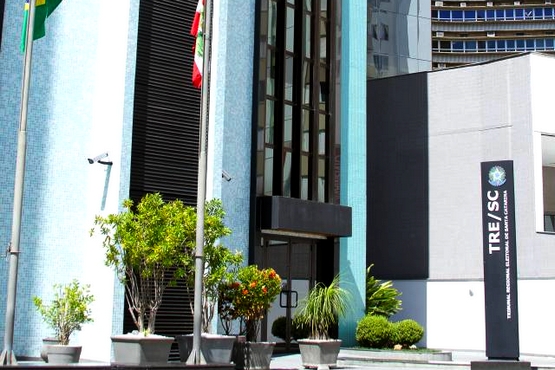 Fachada de um edifício moderno com revestimento em azulejos azuis. Na frente do prédio, há várias plantas em vasos grandes. À esquerda, há mastros com bandeiras, e à direita, uma placa vertical preta com o logotipo do TRE/SC, que significa Tribunal Regional Eleitoral de Santa Catarina. O edifício tem janelas verticais e uma entrada com portas de vidro. Ao fundo, é possível ver parte de outro edifício.