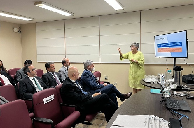 Sala de conferências com várias pessoas sentadas em cadeiras vermelhas, prestando atenção em uma mulher que está em pé, apresentando algo em uma tela de televisão. A mulher está vestida com um vestido verde claro e gesticula com a mão direita enquanto fala. Na mesa em frente a ela, há um laptop, papéis, copos de água e uma garrafa térmica. As cadeiras têm etiquetas com a palavra "RESERVADO". A tela exibe um slide com o título "Sobre o Projeto" e contém texto e gráficos. As pessoas na sala estão vestidas formalmente, com ternos e gravatas. A iluminação é feita por lâmpadas fluorescentes no teto.