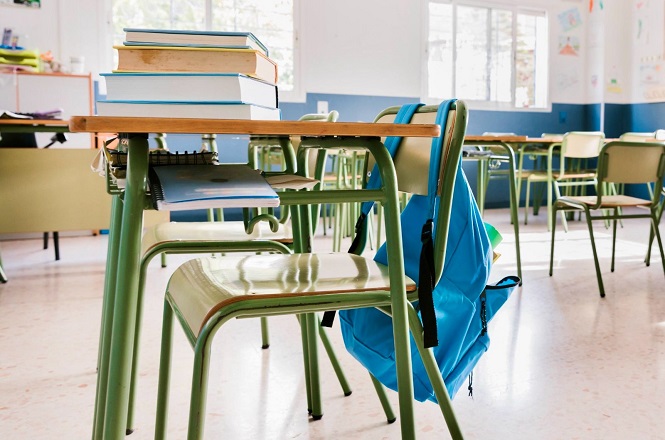Sala de aula com carteiras e cadeiras verdes. Em uma das carteiras, há uma pilha de livros e um caderno. Uma mochila azul está pendurada na cadeira. Ao fundo, é possível ver mais carteiras e cadeiras, além de uma janela grande que permite a entrada de luz natural. As paredes são pintadas de azul na parte inferior e branco na parte superior. Há alguns desenhos pendurados na parede.