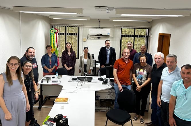 Um grupo de 14 pessoas está reunido em uma sala de conferências. Elas estão posicionadas ao redor de uma mesa em formato de U, que está equipada com computadores, telefones e outros equipamentos de escritório. As pessoas estão sorrindo e olhando para a câmera. A sala tem paredes brancas, duas janelas ao fundo com grades e um aparelho de ar-condicionado acima delas. Há uma decoração colorida pendurada na parede à esquerda. As pessoas estão vestidas de maneira casual e formal, com uma mistura de camisas, blusas e vestidos.