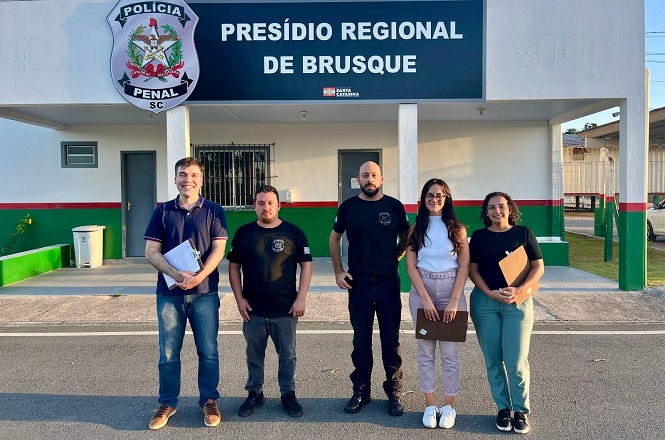 Cinco pessoas estão em pé na frente de um prédio com uma placa que diz "Presídio Regional de Brusque". À esquerda, um homem veste uma camisa polo azul e segura papéis. Ao lado dele, dois homens vestem camisetas pretas com um emblema no peito. À direita, duas mulheres estão lado a lado; uma veste uma blusa branca e calça clara, enquanto a outra usa uma blusa preta e calça jeans. Ambas seguram pastas. O prédio tem uma fachada branca com detalhes em verde e vermelho, e há um emblema da "Polícia Penal SC" à esquerda da placa.