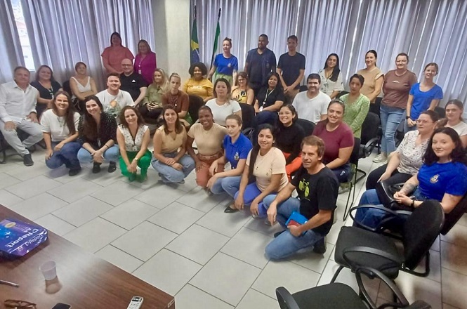 Um grupo de aproximadamente 35 pessoas está reunido em uma sala. Eles estão posando para uma foto em grupo, com algumas pessoas sentadas no chão e outras em pé ou sentadas em cadeiras ao fundo. A maioria das pessoas está sorrindo. No fundo, há cortinas cinzas e duas bandeiras, uma do Brasil e outra que parece ser de um estado ou município. O piso é de azulejos claros e há uma mesa de madeira no canto inferior esquerdo da imagem, sobre a qual há alguns objetos, incluindo um controle remoto. As pessoas estão vestidas de forma casual, com roupas de cores variadas.