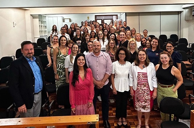 Um grupo grande de pessoas está reunido em uma sala de aula ou auditório. A maioria das pessoas são mulheres, e há um homem no centro do grupo. Todos estão sorrindo e parecem estar participando de um evento ou reunião. As pessoas estão vestidas de maneira casual e formal, com algumas usando vestidos, camisas e calças. As cadeiras da sala estão vazias, sugerindo que o grupo se reuniu na frente para a foto. O ambiente é iluminado e há prateleiras ao fundo.