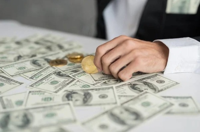 Uma mão masculina segurando moedas douradas sobre uma mesa coberta por várias notas de dólar. A pessoa está vestindo um terno preto com uma camisa branca, e há uma nota de dólar visível no bolso do paletó. As notas de dólar estão espalhadas de forma desordenada pela mesa.