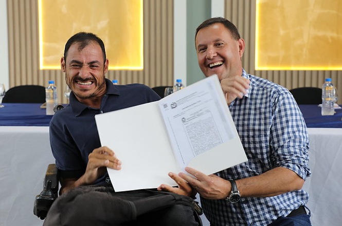 Dois homens estão sorrindo enquanto seguram um documento aberto. O homem à esquerda está em uma cadeira de rodas e veste uma camisa azul escura. O homem à direita veste uma camisa xadrez azul e branca. Ambos parecem estar em um ambiente formal, possivelmente uma cerimônia ou evento, com uma mesa ao fundo coberta por uma toalha azul e garrafas de água sobre ela. As paredes têm detalhes decorativos e iluminação amarela.