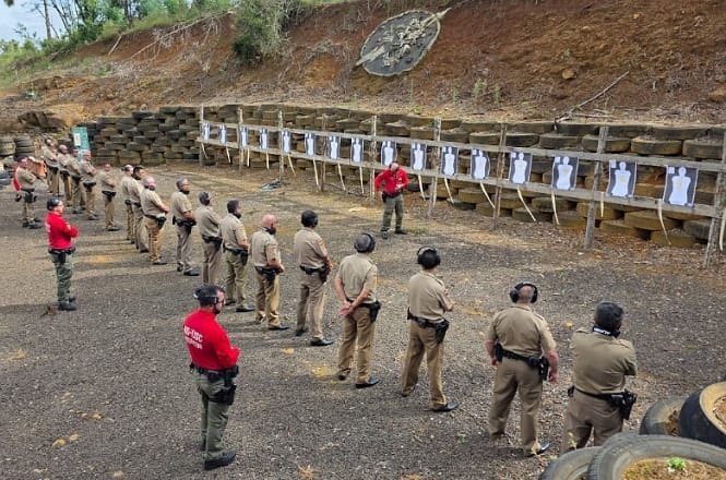Grupo de pessoas em um campo de treinamento de tiro ao ar livre. A maioria está vestida com uniformes marrons e usando protetores auriculares. Eles estão alinhados em frente a alvos de papel que estão presos a uma estrutura de madeira. Dois instrutores, vestidos com camisetas vermelhas, estão na frente do grupo, aparentemente dando instruções. O local é cercado por pneus empilhados e há uma parede de terra ao fundo. A cena sugere uma sessão de treinamento de tiro para forças de segurança.