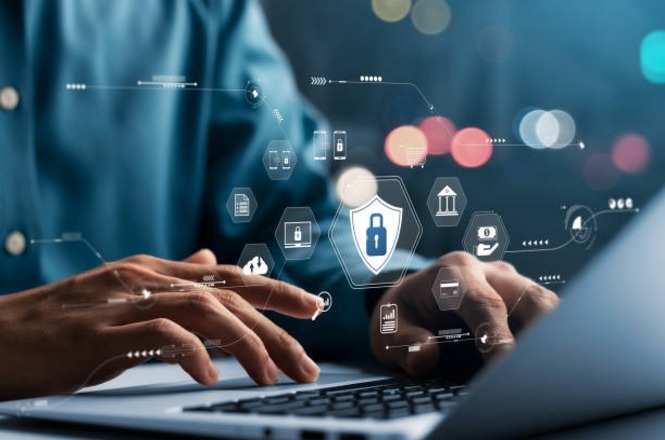 Pessoa digitando em um teclado de laptop. Em torno das mãos, há ícones digitais flutuantes que representam segurança cibernética, como um cadeado em um escudo, além de outros ícones relacionados a tecnologia e segurança. O fundo está desfocado, com luzes coloridas, criando um efeito de profundidade. A cena transmite a ideia de proteção de dados e segurança online.