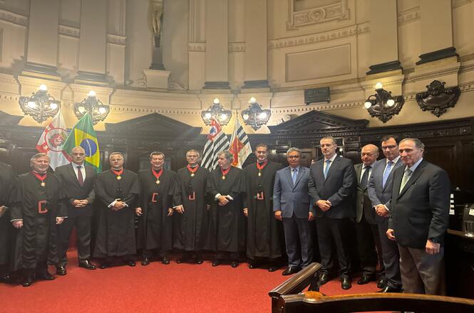 Foto colorida: autoridades alinhadas em plenário histórico, salão solene interno, cerimônia institucional, posando para registro em preto, vermelho e dourado