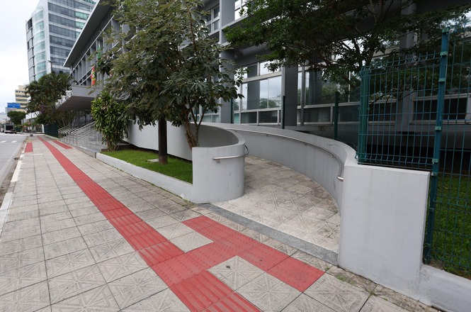 Calçada com piso tátil vermelho para orientação de pessoas com deficiência visual. Ao lado, há uma rampa de acesso com corrimão, cercada por um muro baixo curvo. Árvores e arbustos estão plantados ao longo da rampa. O edifício ao fundo é moderno, com janelas grandes e estrutura de vidro e metal. A rua ao lado é larga e pavimentada, com prédios altos ao fundo.