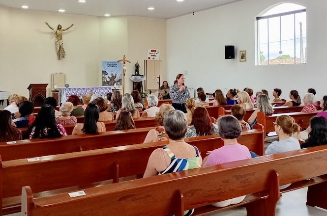 Interior de uma igreja com várias pessoas sentadas em bancos de madeira, voltadas para a frente. A maioria das pessoas são mulheres. Na frente, uma mulher está em pé, aparentemente falando para o grupo. Ao fundo, há um altar com uma estátua de Jesus Cristo na parede. Há também um banner e um microfone próximo ao altar. A iluminação é clara, com luzes no teto e uma janela grande à direita que permite a entrada de luz natural.