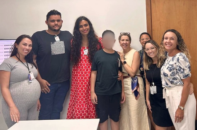 Um grupo de oito pessoas está posando para uma foto em um ambiente interno. À esquerda, há uma mulher grávida vestindo uma camiseta cinza e calças pretas. Ao lado dela, um homem alto com barba está usando uma camiseta preta com um padrão branco no centro e calças jeans. No centro, uma mulher com cabelos longos e cacheados veste um vestido vermelho com estampas brancas. Ao lado dela, um jovem com o rosto desfocado está usando uma camiseta preta e shorts pretos. À direita, uma mulher loira de cabelos presos está usando um vestido bege e óculos de sol no topo da cabeça. Ela está abraçando o jovem. Ao lado dela, uma mulher de óculos com cabelos loiros está usando uma blusa preta e calças escuras. Atrás dela, uma mulher com cabelos presos está parcialmente visíve…