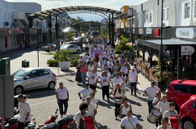Uma multidão de pessoas caminha por uma rua larga e pavimentada. A maioria está vestida com camisetas brancas. Há algumas motos estacionadas à esquerda e à direita da rua. No lado direito, há um estabelecimento com mesas ao ar livre e plantas decorativas. A rua é cercada por prédios comerciais de dois andares. Ao fundo, é possível ver uma estrutura metálica arqueada sobre a rua e algumas árvores. O clima parece ensolarado.