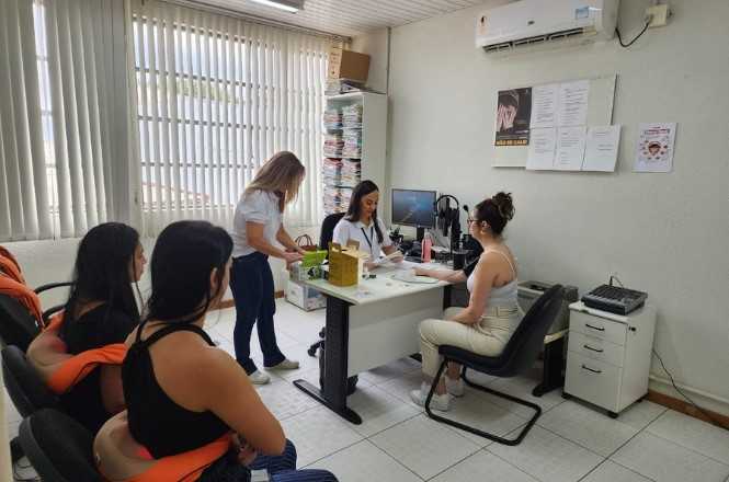 Sala de um consultório ou escritório com quatro mulheres. Duas delas estão sentadas em cadeiras à esquerda, vestindo roupas casuais. Uma terceira mulher está em pé, usando uma camisa branca e calça azul, mexendo em papéis sobre a mesa. A quarta mulher está sentada do outro lado da mesa, de frente para a mulher em pé, usando uma blusa clara e calça branca. A mesa tem vários objetos, incluindo papéis, um computador e uma garrafa de água rosa. Ao fundo, há uma estante cheia de papéis e um ar-condicionado na parede. Há também cartazes e papéis fixados na parede. A sala é iluminada por luz natural que entra através de persianas na janela.